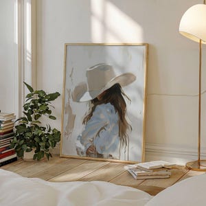 Pode incluir: Uma impress&atilde;o de arte emoldurada retrata uma pessoa usando um chap&eacute;u de cowboy e uma camisa azul clara. A obra de arte tem uma paleta de cores neutras com uma moldura dourada. A pintura est&aacute; encostada em uma parede branca, com uma planta e livros ao fundo.