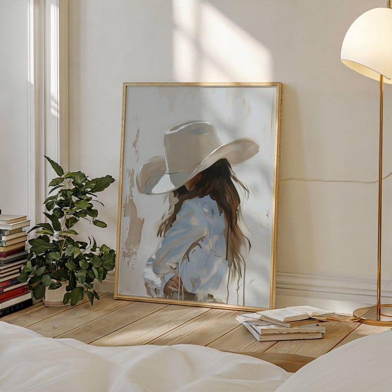 Pode incluir: Uma impress&atilde;o de arte emoldurada retrata uma pessoa usando um chap&eacute;u de cowboy e uma camisa azul clara. A obra de arte tem uma paleta de cores neutras com uma moldura dourada. A pintura est&aacute; encostada em uma parede branca, com uma planta e livros ao fundo.