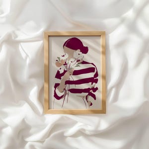 May include: A framed wall art print featuring a stylized illustration of a person with dark hair wearing a white shirt with horizontal maroon stripes. The person is holding a small bouquet of white flowers near their face against a light background.