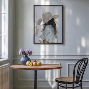 Pode incluir: Uma obra de arte emoldurada retrata uma pessoa usando um chap&eacute;u de cowboy branco e uma camisa azul clara. A pintura est&aacute; pendurada acima de uma mesa redonda de madeira com uma base de metal preta. Um vaso azul com flores roxas e ma&ccedil;&atilde;s est&aacute; sobre a mesa. Uma cadeira preta est&aacute; por perto.