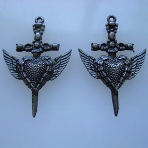 May include: Two silver-toned metal charms in the shape of a heart pierced by a dagger with wings. The heart is textured and the dagger has a cross at the top.