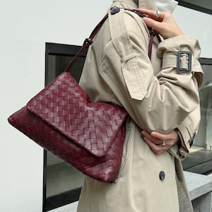 May include: A deep burgundy shoulder bag featuring a woven, grid-like pattern across its surface. The bag has a fold-over flap closure and a thin, adjustable strap that connects to the sides, creating a structured yet flexible silhouette.