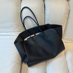 May include: A black leather tote bag with long handles. The bag has a rectangular shape with a wide opening and a smooth, matte finish. The bag is sitting on a white surface.