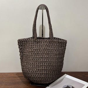Può includere: Una borsa tote marrone scuro caratterizzata da una lavorazione all'uncinetto a maglia aperta. La borsa presenta due manici abbinati fissati al bordo superiore, un'ampia apertura e una base arrotondata con un motivo a punti ripetuto.