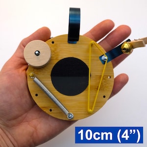 May include: A circular wooden toy, approximately 10cm (4") in diameter, featuring a black central section, a spring, and a yellow elastic band. It includes a wooden wheel and a blue metal component. The text "10cm (4")" is displayed.