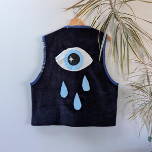 Pode incluir: Colete de veludo cotelê azul marinho com um grande design de olho e lágrimas. O olho é branco com uma íris azul claro e uma pupila preta com uma estrela. O colete tem uma borda azul e está pendurado em um cabide de madeira.