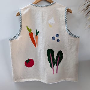 Op de afbeelding: Een mouwloos vest in off-white met kleurrijke stoffen groente-applicaties, waaronder wortels, een tomaat, bosbessen, knoflook en bladgroenten. Het kledingstuk is afgewerkt met een lichtblauw en wit gingham-biesje langs de armsgaten en halslijn.