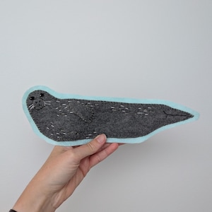 Peut inclure: Un ornement plat en feutrine en forme de phoque gris foncé avec une bordure bleu clair. Le phoque présente des yeux en perles noires, des moustaches blanches brodées et des détails cousus blancs sur le corps, les nageoires et la queue.