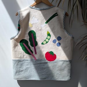 May include: A sleeveless waistcoat in off-white fabric featuring colourful fabric appliqués of vegetables including chard, a pea pod, a tomato, garlic, and a spring onion. The waistcoat has a grey and white gingham trim along the edges and a bottom panel.