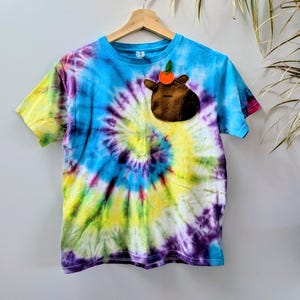 May include: A colourful tie-dye t-shirt in blue, yellow, and purple, featuring a brown felt applique of a cartoon animal with an orange fruit. The shirt is displayed on a wooden hanger against a white wall.