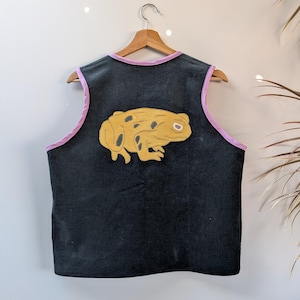 Puede incluir: Un chaleco de pana negro que presenta un sapo grande, amarillo y bordado con manchas oscuras en la parte posterior. El chaleco está rematado con un ribete de color violeta claro a lo largo del cuello y las sisas, y se muestra en una percha de madera.