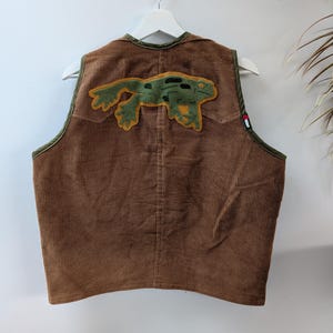 May include: A brown corduroy vest featuring a large green frog appliqué on the back. The vest has olive green trim along the armholes and neckline, and a small rectangular tag with a red and white graphic on the side seam.