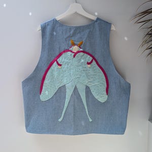 May include: A light blue denim waistcoat featuring a large, central appliqué of a moth. The moth has pale green wings with dark pink borders, a cream-coloured body, and orange antennae. The waistcoat is sleeveless with a round neckline and hangs on a white hanger.