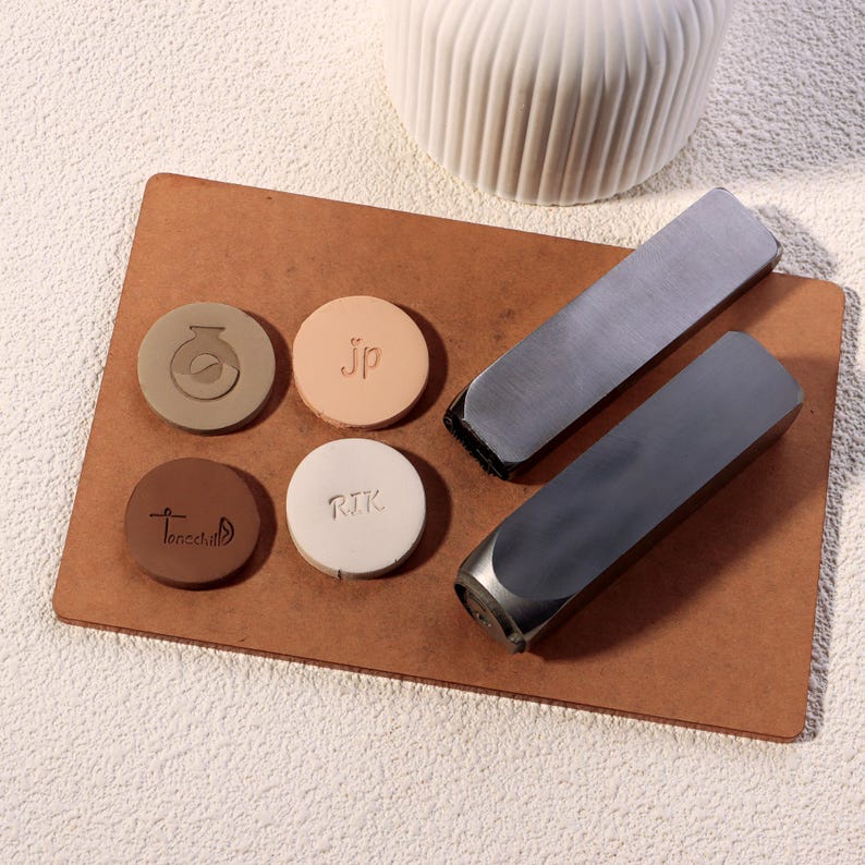 May include: A collection of personalized leather stamps and metal tools arranged on a brown wooden board. The stamps are circular, in various colors, and feature initials and designs. Two metal tools with black edges are also present.