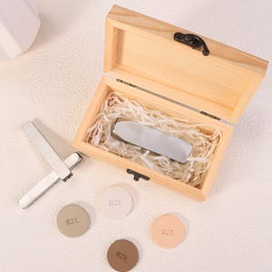 May include: A wooden box with a hinged lid, containing a silver metal object nestled in paper-like filler. Several small, round, leather-like pieces with the letters "RIK" are scattered around the box, along with two silver metal tools.