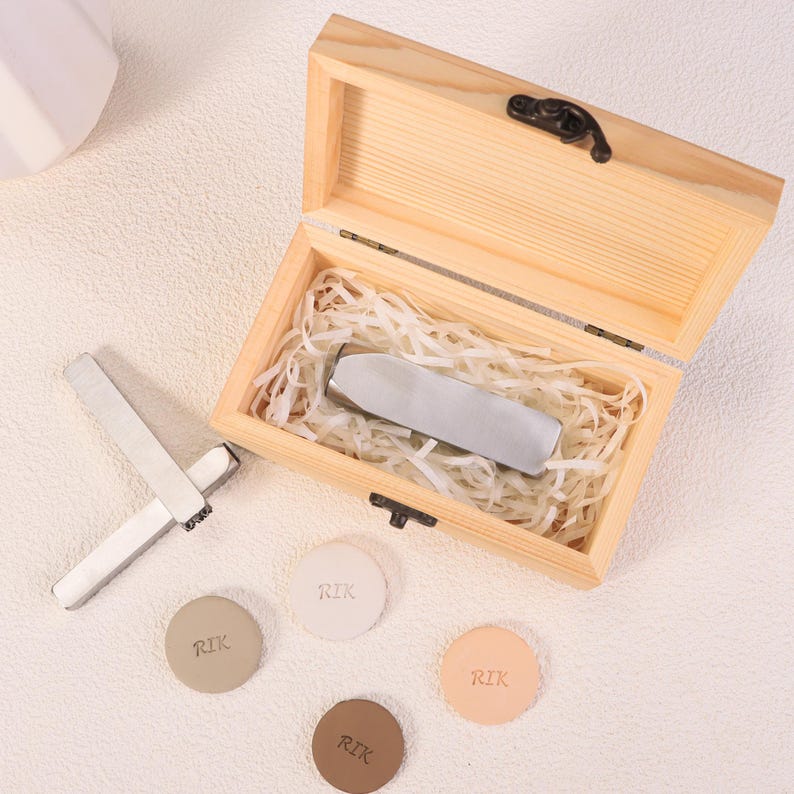 May include: A wooden box with a hinged lid, containing a silver metal object nestled in paper-like filler. Several small, round, leather-like pieces with the letters "RIK" are scattered around the box, along with two silver metal tools.