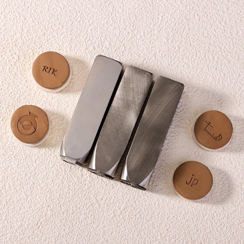 May include: An overhead shot shows three metal stamps and four small, round, brown objects with white rims. The stamps are rectangular with a beveled edge. The brown objects have text and a graphic design.