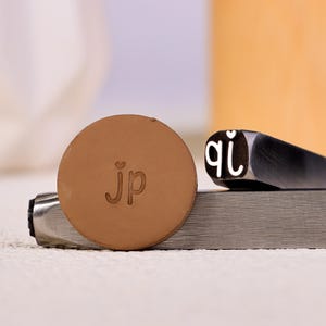 May include: Close-up of metal letter stamps and a round, brown leather piece with the initials "jp" stamped on it. The stamps are used for imprinting letters and designs. The background is blurred, with a neutral color palette.