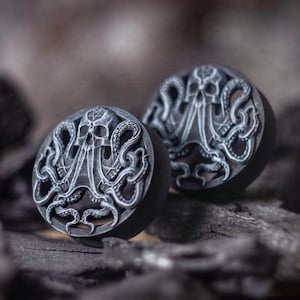 Può includere: Un paio di dilatatori per orecchie in legno nero con una scultura dettagliata di un polpo con un teschio al centro.