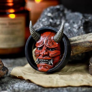 Può includere: Una maschera Oni rossa e nera con corna, inserita in una cornice circolare nera. La maschera ha un'espressione feroce con denti aguzzi e occhi bianchi. La maschera è su una superficie marrone.