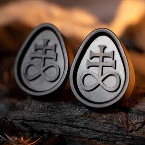 May include: Two dark gray, teardrop-shaped plugs with the symbol of Leviathan. The plugs are made of a dark gray material and have a raised design. The background is a dark, textured surface.