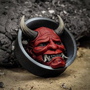 May include: A red and black Oni mask with horns, set in a black circular frame. The mask has a fierce expression with sharp teeth and white eyes.