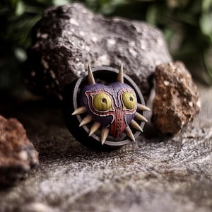 Può includere: Un primo piano di una replica della Maschera di Majora. La maschera è realizzata in legno dipinto e presenta una combinazione di colori viola scuro, rosso e giallo. La maschera ha la forma di un cuore ed è circondata da un anello nero con punte.
