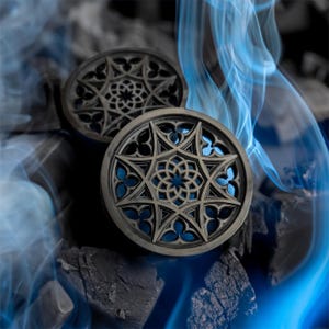 May include: Two black wooden ear gauges with a geometric design resembling a stained glass window. The gauges are set against a background of black charcoal and blue smoke.