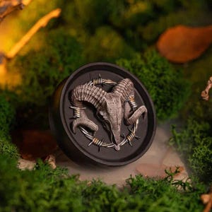 Pode incluir: Um objeto redondo preto com um design detalhado de crânio de carneiro. O crânio é cinza com chifres curvos, cercado por arame farpado. O fundo é uma mistura desfocada de verde e marrom.