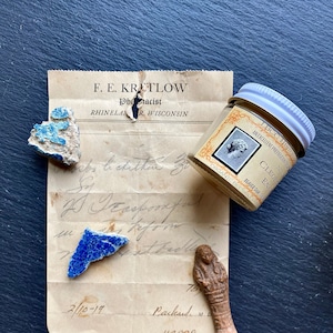 May include: A small glass jar with a white lid containing a light brown substance. The jar has a label that reads "LBCC Historical Apothecary No. CLEOPATRA ENAMEL BEAVER DAM ORGANIC BEESWAX MIRACLE". The jar is sitting on a piece of paper with handwritten text. There are also three small pieces of blue and green tile and a small brown figurine.