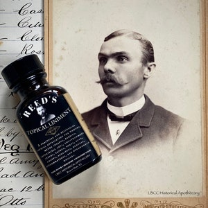 May include: A vintage photo of a man with a mustache, wearing a suit and bow tie. The photo is mounted on a card with the text "LBCC Historical Apothecary" and "Stevens Chicago Extra Finish" printed on it. A small brown glass bottle with a black cap and the text "Reed's Topical Liniment" is in the foreground.
