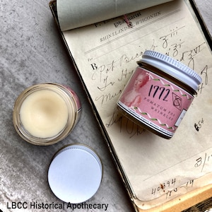 May include: A vintage apothecary jar with a white lid and a pink label that reads "1772 Pomatum for Wrinkles". The jar is sitting on a page from an old apothecary notebook with handwritten text.
