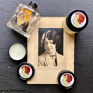 May include: A vintage photo of a woman with dark hair and a white blouse is displayed on a brown paper background.  Four small glass jars with black lids are arranged around the photo.  The jars are labeled "Petroleum" and feature a vintage illustration of a woman with red hair.  A clear glass bottle with a black cap is also in the image.  The bottle is labeled "Star Black Brilliantine" and features a vintage illustration of a man.