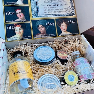 May include: A gift box containing historical-style apothecary products nestled in wood shavings. The lid features portraits and text including Care of the Skin, Cosmetics, and dates from 1803 to 1829. Items include glass bottles and jars labeled Orange Flower Water, Milk of Roses, and Pearl Powder.