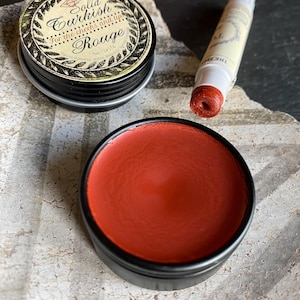 Könnte beinhalten: Ein fester Rouge für Wangen und Lippen in einer schwarzen Dose, mit einem passenden Lippenbalsam. Der Rouge hat eine tiefrote Farbe. Auf der Dose steht "Solid Turkish Rouge". Der Lippenbalsam befindet sich in einer weißen Tube.