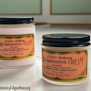 May include: Two small jars of LBCC Apothecary Flesh Vanishing Cream, with black lids and labels featuring text. The labels read "Rose Healing and Soothing" and "Beaver Dam, WI". The jars are on a white surface.