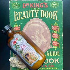 May include: An antique brown glass bottle of "Bath Crystals Rose Geranium" rests on a vintage green "Dr. King's New Beauty Book." The bottle has a decorative label with floral and portrait illustrations. The book's cover features text and illustrations.