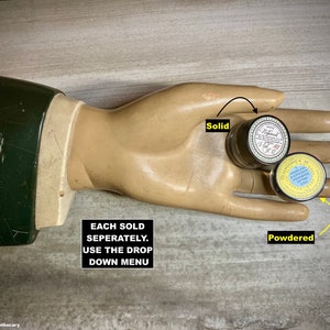 May include: Two small, round tins labeled "Solid" and "Powdered" rest on a mannequin hand. The tins have white and yellow labels with black text. The mannequin hand is beige with a green-sleeved arm.