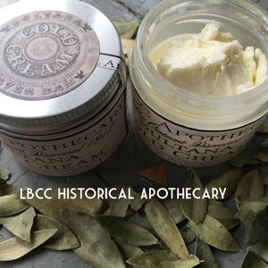 K&ouml;nnte beinhalten: Zwei kleine Cremegl&auml;ser, eines mit einem silbernen Deckel und das andere mit einem durchsichtigen Deckel. Der silberne Deckel hat ein Etikett mit der Aufschrift "Gold Cream". Der durchsichtige Deckel hat ein Etikett mit der Aufschrift "Sultanana Cream". Beide Gl&auml;ser stehen auf einem Bett aus getrockneten Bl&auml;ttern.