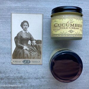 May include: A vintage photo of a woman in a plaid dress, sitting in a chair. The photo is framed and has a white background. The photo is next to a jar of cucumber cold cream with a gold lid and label. The label reads "Cucumber Cold Cream" and "The 1857" in a vintage font.