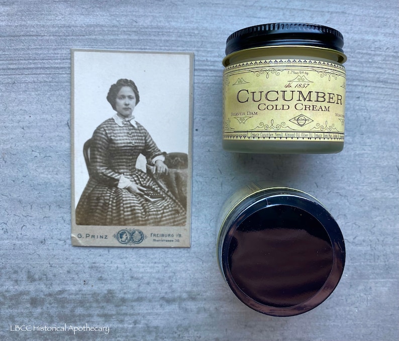 May include: A vintage photo of a woman in a plaid dress, sitting in a chair. The photo is framed and has a white background. The photo is next to a jar of cucumber cold cream with a gold lid and label. The label reads "Cucumber Cold Cream" and "The 1857" in a vintage font.