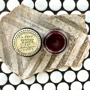 May include: A small jar of tinted lip salve with a black lid and a gold label that reads "A FINE TINTED LIP SALVE An original & Harmless Preparation 1772". The jar is sitting on a piece of broken pottery.
