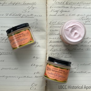 May include: Three jars of LBCC Apothecary Flesh Vanishing Cream with Rose, a healing and soothing cream. The jars have black lids and labels with text. The cream is pink and sits on a vintage ledger.