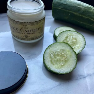 May include: A glass jar of cucumber cold cream with a gold label that says "Cucumber Cold Cream" and "Est. 1857". The jar is sitting on a white marble surface with several slices of cucumber beside it.