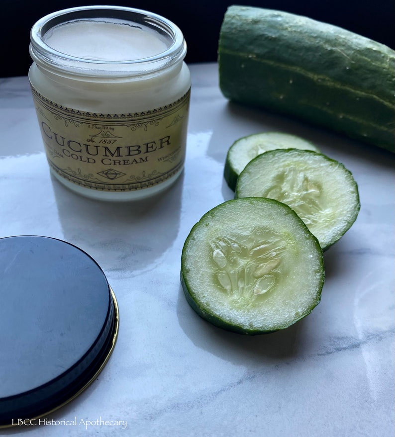May include: A glass jar of cucumber cold cream with a gold label that says "Cucumber Cold Cream" and "Est. 1857". The jar is sitting on a white marble surface with several slices of cucumber beside it.