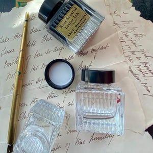 May include: Two glass ink bottles with black lids and a gold-colored dip pen on a piece of paper with handwritten text. The ink bottles are labeled "Rose Scented Letter Ink" and "LBCC Historical Apothecary".