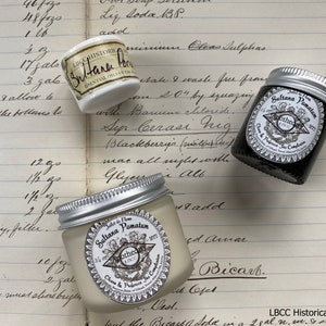 May include: Three small jars of cream or balm with silver lids. The jars are labeled "Sultana Pomatum" and "Sultana Balm" with the text "Apothecary London" on the labels. The jars are sitting on a page of a notebook with handwritten text.