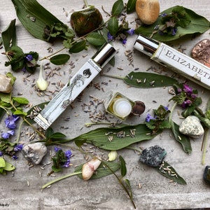May include: Two glass bottles of perfume with silver caps, one with a label that says "Elizabeth", surrounded by green leaves, purple flowers, and various stones.