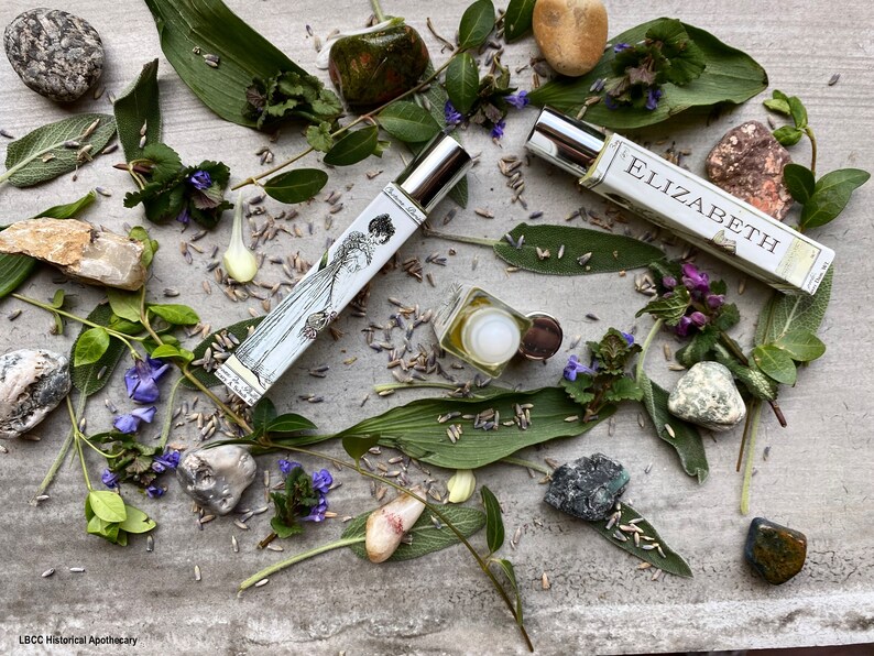 May include: Two glass bottles of perfume with silver caps, one with a label that says "Elizabeth", surrounded by green leaves, purple flowers, and various stones.
