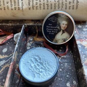 May include: Two small, round metal tins. One tin has a label with a portrait and the text "AN EXCELLENT PAINT FOR THE FACE .5 OZ". The other tin is open, revealing a white, textured substance. The tins are in a decorative box.